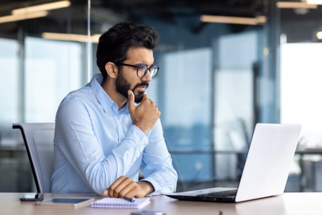 An investor sitting at a desk, thinking as they look at a laptop.
