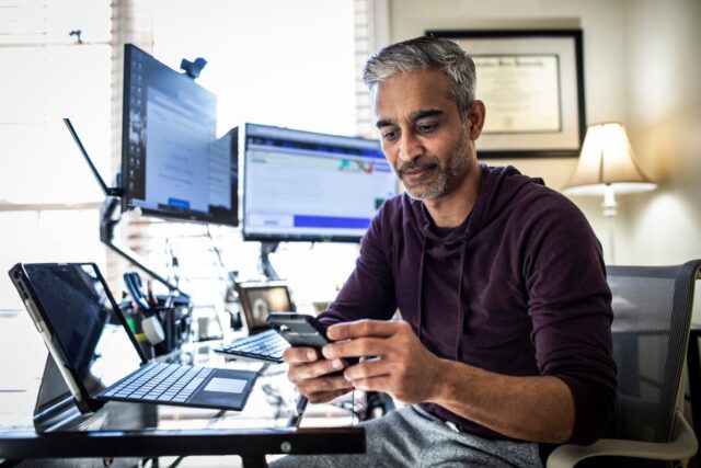 A man trading stocks on his phone at his desk with a lot of computers around him. 